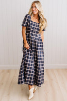 A woman modeling a navy and white plaid midi dress with short puff sleeves, a button-front bodice, and side pockets, paired with white cowboy boots, standing against a white paneled wall and light wood floor.