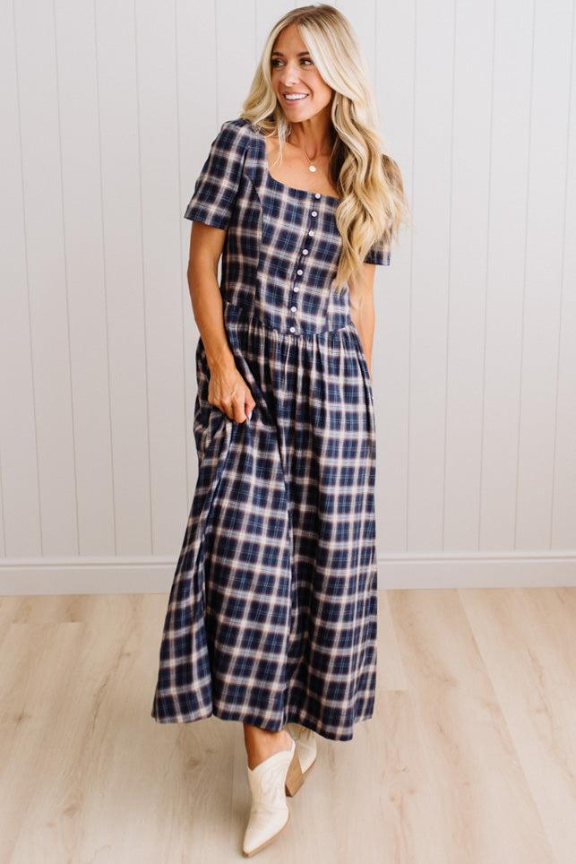 A woman modeling a navy and white plaid midi dress with short puff sleeves, a button-front bodice, and side pockets, paired with white cowboy boots, standing against a white paneled wall and light wood floor.
