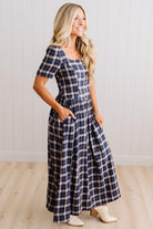 A woman modeling a navy and beige plaid short-sleeve maxi dress with pockets, standing in a room with light wood floors and white paneled walls.