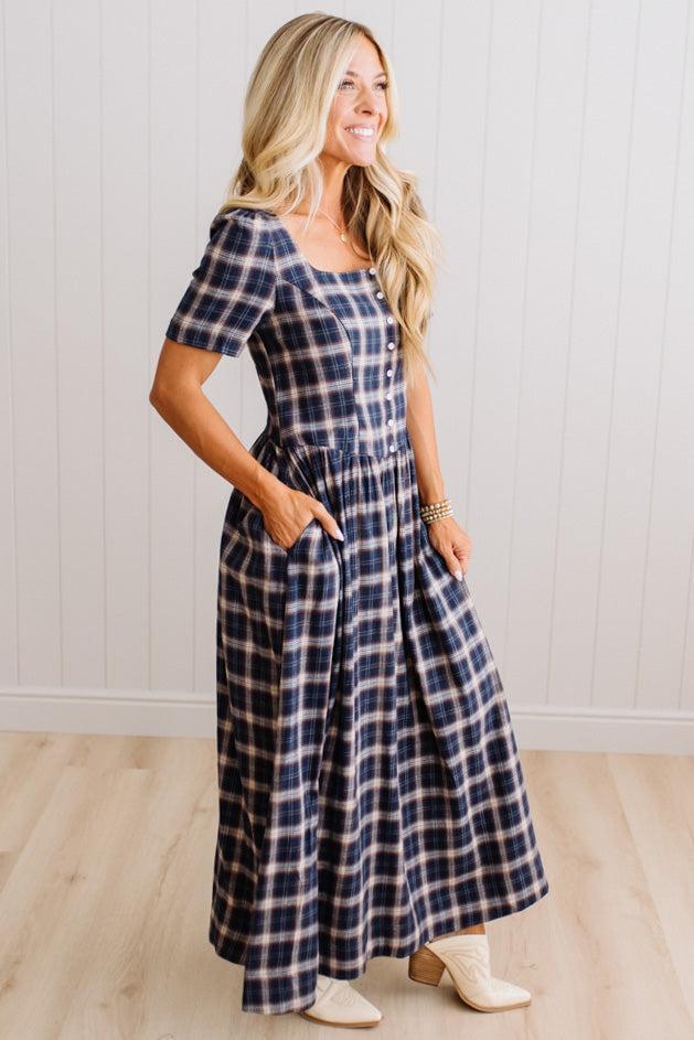 A woman modeling a navy and beige plaid short-sleeve maxi dress with pockets, standing in a room with light wood floors and white paneled walls.