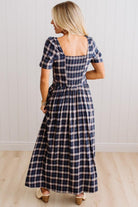 A woman wearing a navy blue and beige plaid midi dress, shown from the back against a white paneled wall and light wood floor.