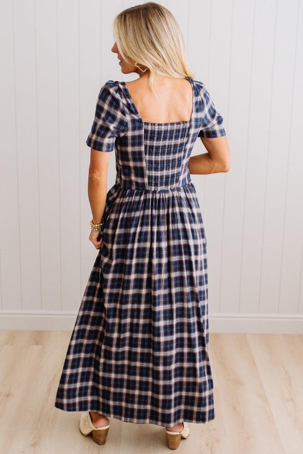 A woman wearing a navy blue and beige plaid midi dress, shown from the back against a white paneled wall and light wood floor.