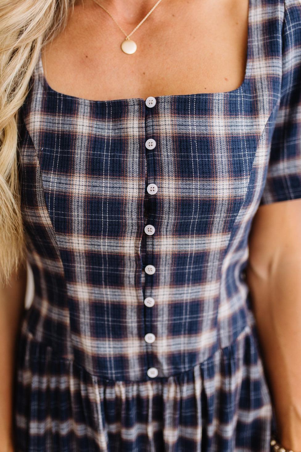 Close-up of a woman wearing a navy blue plaid dress with white and light brown stripes, featuring a square neckline and a button-down front.