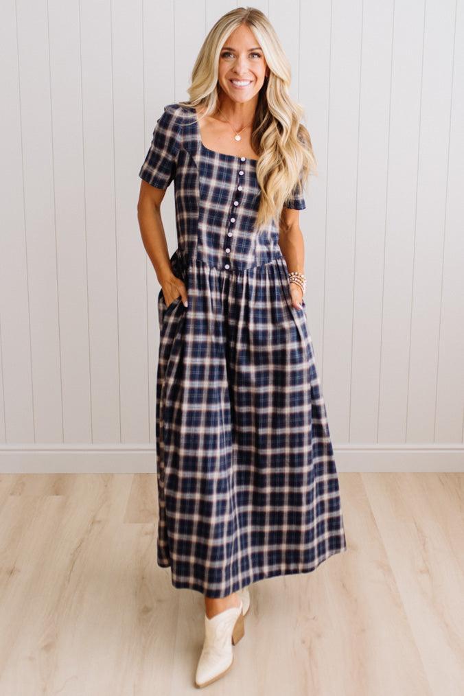 A woman modeling a navy and white plaid midi dress with short sleeves, a square neckline, and a button-up front, standing in front of a white paneled wall and light wood floor.
