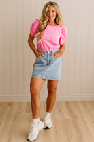 Woman wearing a pink top and denim skirt standing against a beige wall.