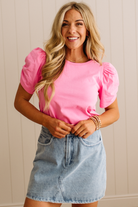 Woman wearing a pink top with puff sleeves and a denim skirt against a beige wall.