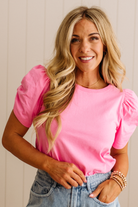Woman wearing a pink top with puffed sleeves and denim shorts against a neutral background