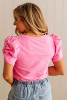 Woman wearing a pink top with ruffled sleeves and light blue jeans against a white wall.