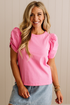 Woman wearing a pink top with puff sleeves and denim shorts against a beige wall.