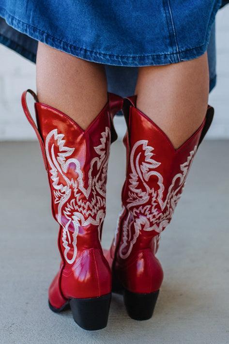 Red cowboy boots with white patterns worn by a person in a blue skirt.