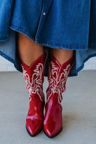 Red cowboy boots with white patterns worn with a blue denim outfit on a light background