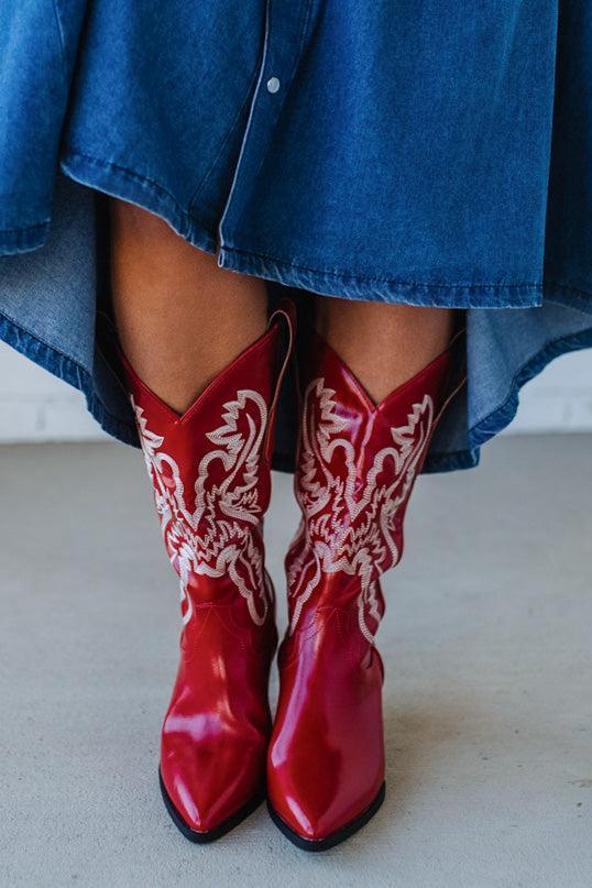 Red cowboy boots with white patterns worn with a blue denim outfit on a light background