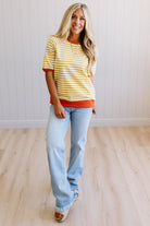 Blonde woman wearing light jeans and a mustard yellow with white stiripe short sleeve top, that also has a dark orange contrast trim.