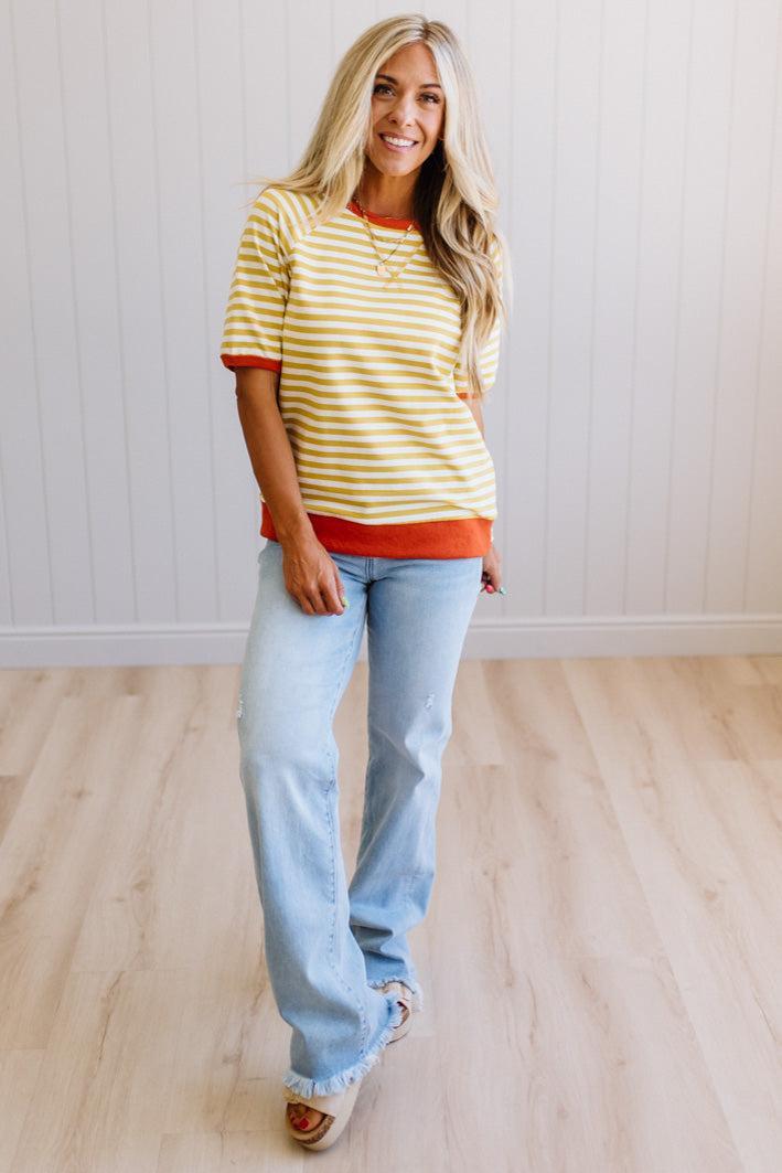 Blonde woman wearing light jeans and a mustard yellow with white stiripe short sleeve top, that also has a dark orange contrast trim.