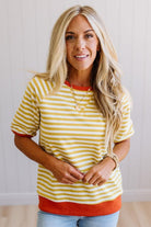 Blonde woman wearing light jeans and a mustard yellow with white stiripe short sleeve top, that also has a dark orange contrast trim.