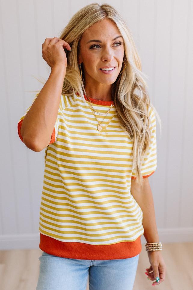 Blonde woman wearing light jeans and a mustard yellow with white stiripe short sleeve top, that also has a dark orange contrast trim.