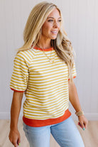 Blonde woman wearing light jeans and a mustard yellow with white stiripe short sleeve top, that also has a dark orange contrast trim.
