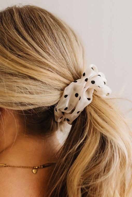 A close-up of a blonde woman's ponytail secured with a cream-colored fabric scrunchie featuring black polka dots, shown from the side against a light neutral background.