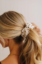 A blonde woman shown from the back wearing a white fabric scrunchie with black polka dots in her ponytail, against a plain light background.
