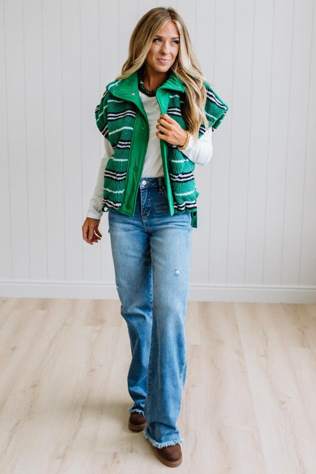 BLONDE WOMAN IN A STUDIO WITH LIGHT WOOD FLOORS AND LIGHT BACKDROP WEARING BLUE JEANS, A LONG SLEEVE WHITE TEE SHIRT AND A GREEN BLACK AND WHITE SHERPA COMFORT STRIPED VEST WITH SNAP BUTTONS AND SIDE POCKETS