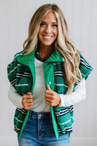 BLONDE WOMAN IN A STUDIO WITH LIGHT WOOD FLOORS AND LIGHT BACKDROP WEARING BLUE JEANS, A LONG SLEEVE WHITE TEE SHIRT AND A GREEN BLACK AND WHITE SHERPA COMFORT STRIPED VEST WITH SNAP BUTTONS AND SIDE POCKETS