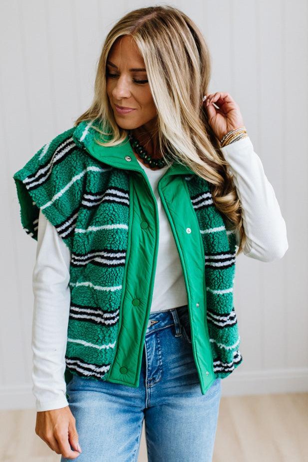 BLONDE WOMAN IN A STUDIO WITH LIGHT WOOD FLOORS AND LIGHT BACKDROP WEARING BLUE JEANS, A LONG SLEEVE WHITE TEE SHIRT AND A GREEN BLACK AND WHITE SHERPA COMFORT STRIPED VEST WITH SNAP BUTTONS AND SIDE POCKETS