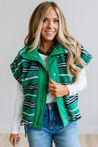 BLONDE WOMAN IN A STUDIO WITH LIGHT WOOD FLOORS AND LIGHT BACKDROP WEARING BLUE JEANS, A LONG SLEEVE WHITE TEE SHIRT AND A GREEN BLACK AND WHITE SHERPA COMFORT STRIPED VEST WITH SNAP BUTTONS AND SIDE POCKETS