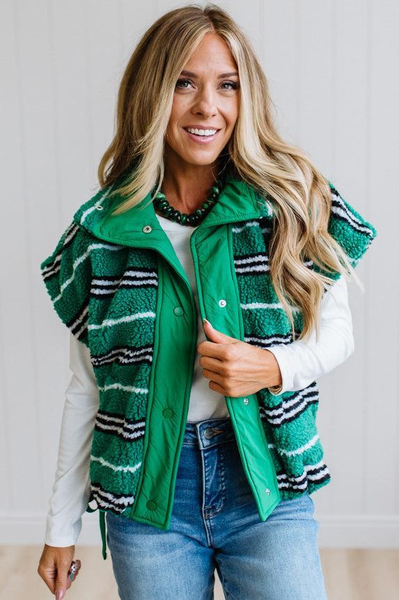 BLONDE WOMAN IN A STUDIO WITH LIGHT WOOD FLOORS AND LIGHT BACKDROP WEARING BLUE JEANS, A LONG SLEEVE WHITE TEE SHIRT AND A GREEN BLACK AND WHITE SHERPA COMFORT STRIPED VEST WITH SNAP BUTTONS AND SIDE POCKETS
