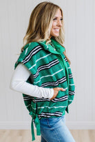 BLONDE WOMAN IN A STUDIO WITH LIGHT WOOD FLOORS AND LIGHT BACKDROP WEARING BLUE JEANS, A LONG SLEEVE WHITE TEE SHIRT AND A GREEN BLACK AND WHITE SHERPA COMFORT STRIPED VEST WITH SNAP BUTTONS AND SIDE POCKETS