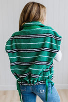 BLONDE WOMAN IN A STUDIO WITH LIGHT WOOD FLOORS AND LIGHT BACKDROP WEARING BLUE JEANS, A LONG SLEEVE WHITE TEE SHIRT AND A GREEN BLACK AND WHITE SHERPA COMFORT STRIPED VEST WITH SNAP BUTTONS AND SIDE POCKETS