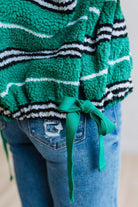 BLONDE WOMAN IN A STUDIO WITH LIGHT WOOD FLOORS AND LIGHT BACKDROP WEARING BLUE JEANS, A LONG SLEEVE WHITE TEE SHIRT AND A GREEN BLACK AND WHITE SHERPA COMFORT STRIPED VEST WITH SNAP BUTTONS AND SIDE POCKETS