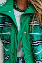 A close-up of a person wearing a bright green sherpa-lined puffer jacket with green, black, and white striped fleece panels, layered over a white top and blue jeans.