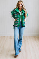 BLONDE WOMAN IN A STUDIO WITH LIGHT WOOD FLOORS AND LIGHT BACKDROP WEARING BLUE JEANS, A LONG SLEEVE WHITE TEE SHIRT AND A GREEN BLACK AND WHITE SHERPA COMFORT STRIPED VEST WITH SNAP BUTTONS AND SIDE POCKETS