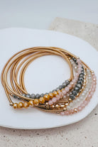A stack of gold-toned beaded stretch bracelets arranged on a white ceramic plate. The bracelets feature faceted beads in gold, amber, gray, and soft pink, with small metallic gold spacer beads.