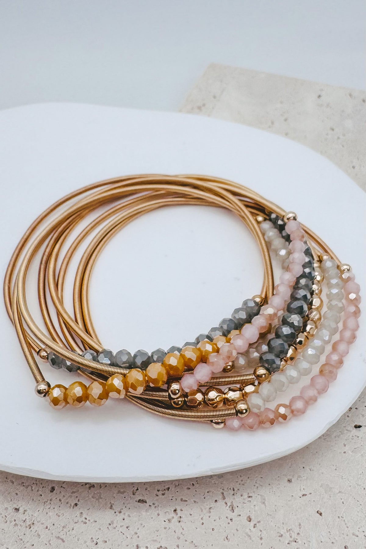 A stack of gold-toned beaded stretch bracelets arranged on a white ceramic plate. The bracelets feature faceted beads in gold, amber, gray, and soft pink, with small metallic gold spacer beads.