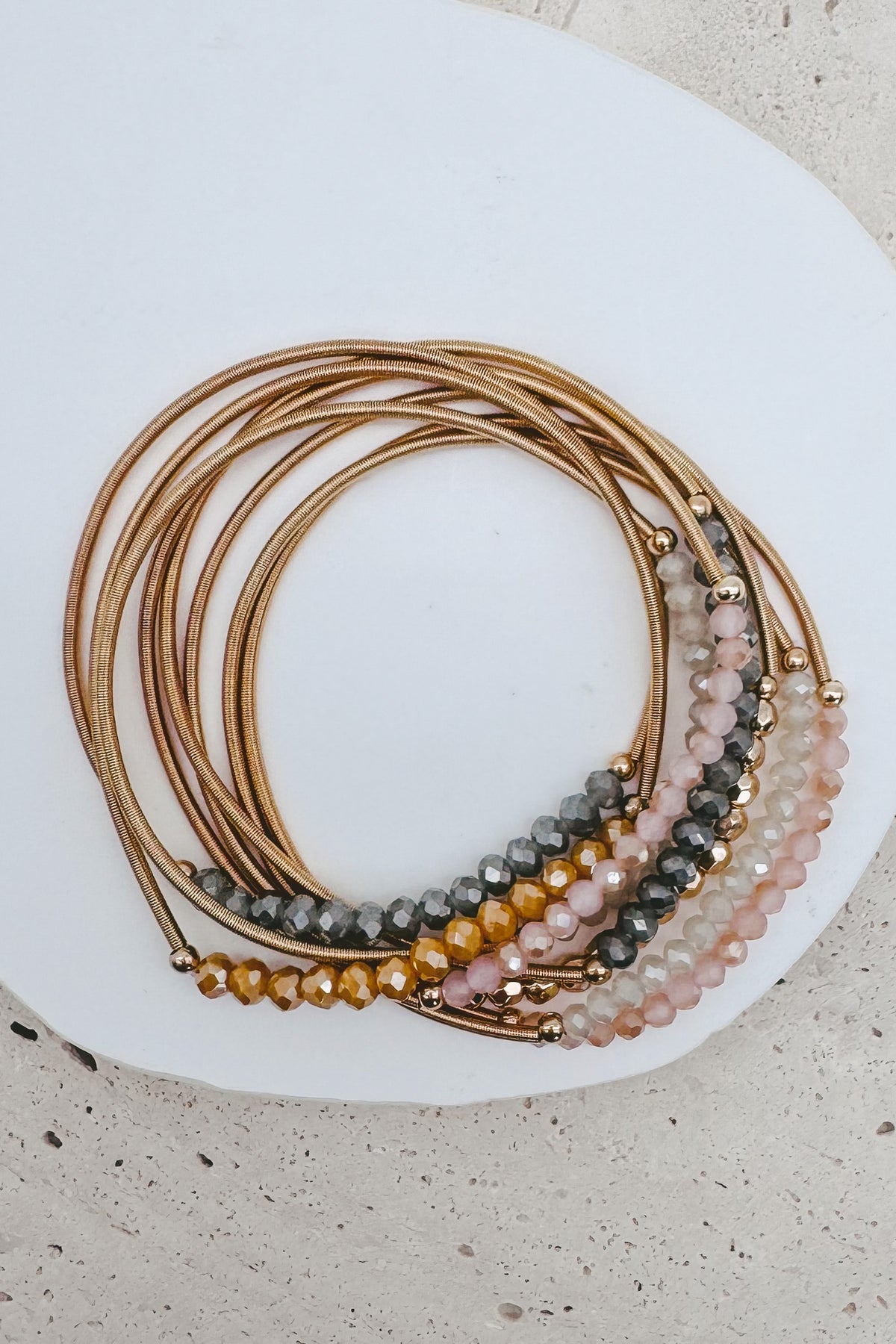 A stack of multi-strand beaded bracelets with gold-toned metal bands, displayed on a white ceramic plate over a light beige textured surface.