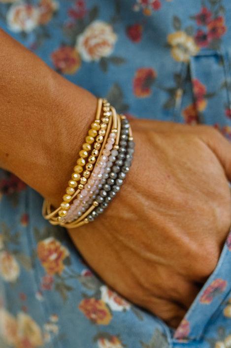 A close-up of a person's wrist wearing a stack of beaded bracelets, including gold, light pink, and gray beads, against a blue floral shirt background.