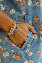 A close-up of a person's hand in a floral blue pocket, wearing a stack of beaded wrap bracelets in gold, silver, and rose gold tones.