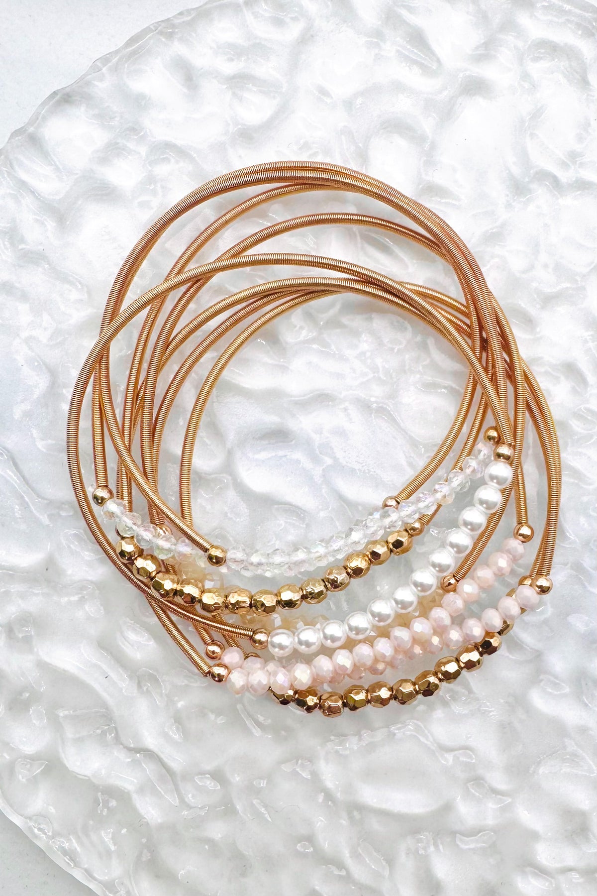 A close-up of a stack of gold and pearl beaded bracelets, on a textured surface.