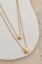 Two gold heart-shaped pendants on delicate chains against a neutral background