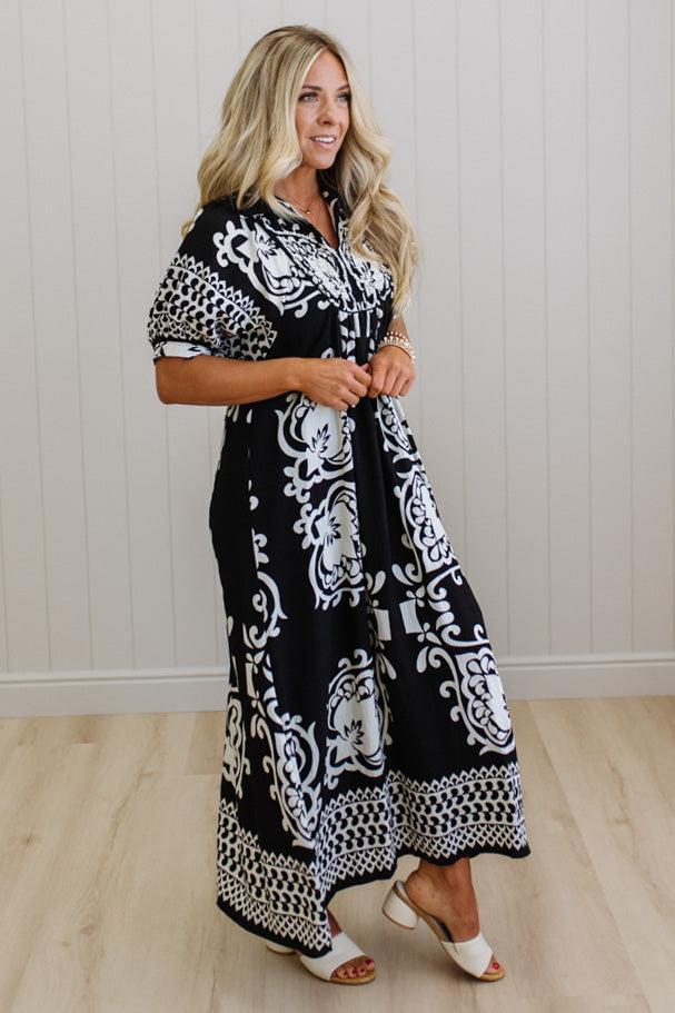 Woman wearing a black and white patterned dress standing against a white wall.