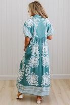 Woman wearing a teal dress with white patterns in a room with a light wooden floor and white paneled walls.