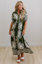Woman wearing a green and white patterned dress against a white paneled wall.