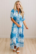 A woman modeling a blue and white patterned maxi dress with short sleeves and a V-neckline, standing against a white paneled wall and light wood floor.