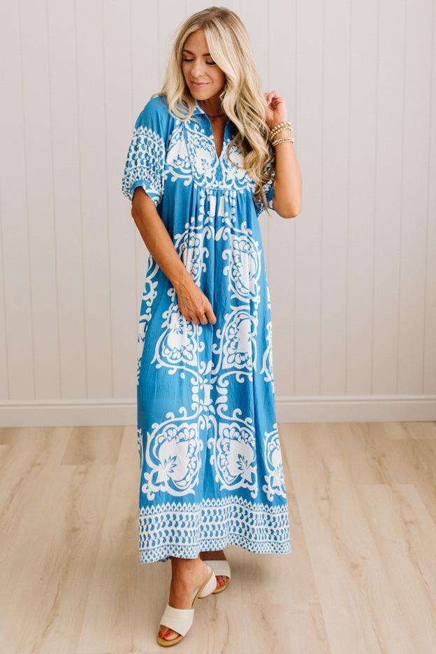 A woman modeling a blue and white patterned maxi dress with short sleeves and a button-up front, standing in a room with light wood floors and a white paneled wall.