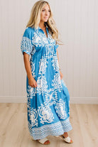 A woman modeling a blue and white patterned maxi dress with short puff sleeves, a V-neckline, and a tie at the chest, standing in a room with light wood floors and white paneled walls.