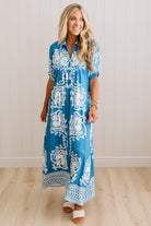 A woman modeling a blue and white patterned maxi dress with short sleeves, a tie neckline, and a loose, flowy silhouette, standing in a bright indoor setting with light wood floors and white paneled walls.