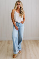 Woman wearing a white ribbed tank top and light blue jeans standing against a white wall.