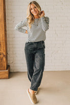 Woman wearing a striped long-sleeve shirt and dark jeans standing against a white brick wall.