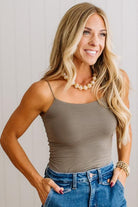 A smiling blonde woman wearing a fitted taupe-brown spaghetti-strap camisole top and blue jeans, standing against a light neutral paneled wall.